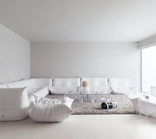 Soggiorno minimalista con divano componibile bianco, tappeto grigio e tavolino trasparente. Le tonalità di bianco per pareti creano un'atmosfera eterea.